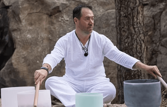 A practitioner seated outdoors in white clothing, playing a set of crystal singing bowls.