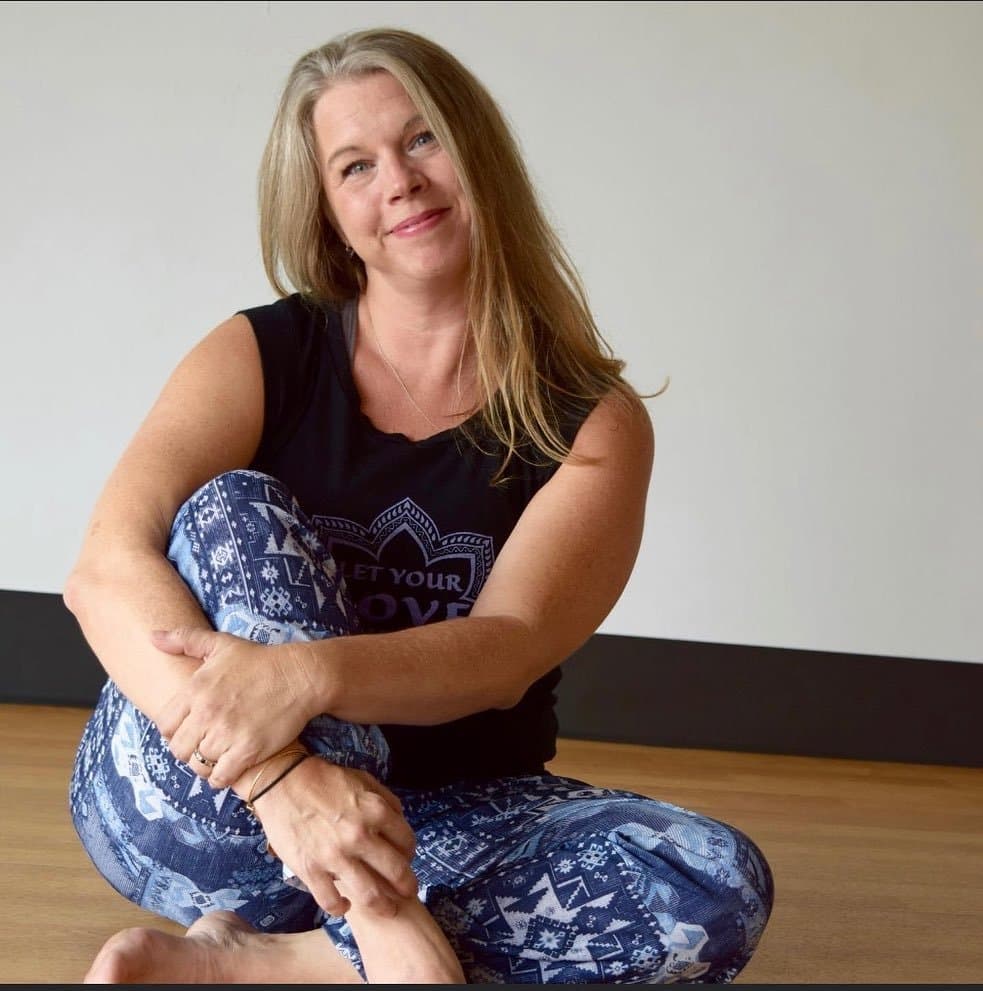 Beth Evans, yoga therapist, sitting cross-legged on a wooden floor and smiling gently.